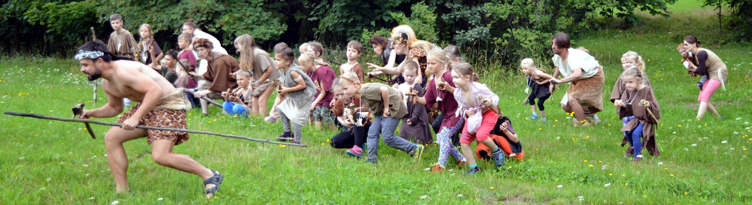 Děti oblečené do kostýmů pravěkých lidí se v přírodě plíží při lovu mamuta